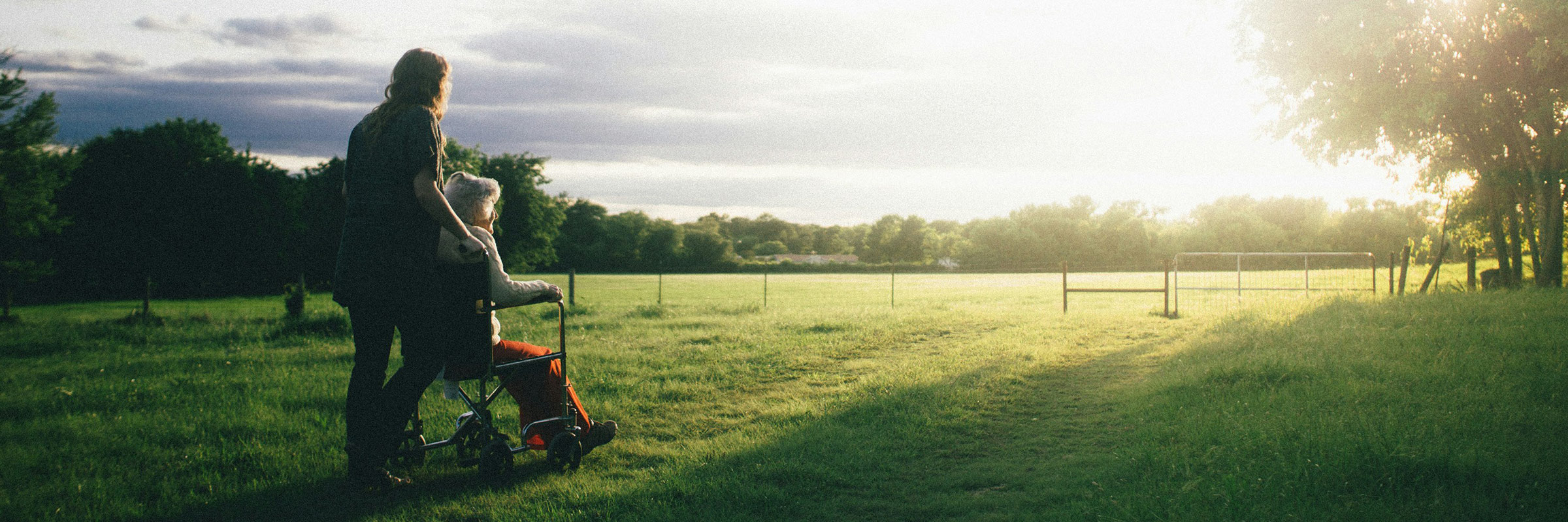 long-haired person pushing elderly person in wheelchair through field towards sunset