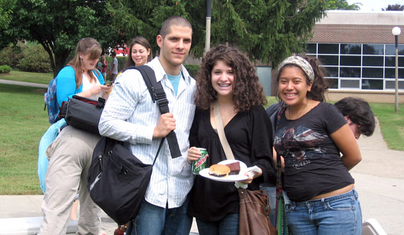 three students smiling outside on campus. one is holding food.